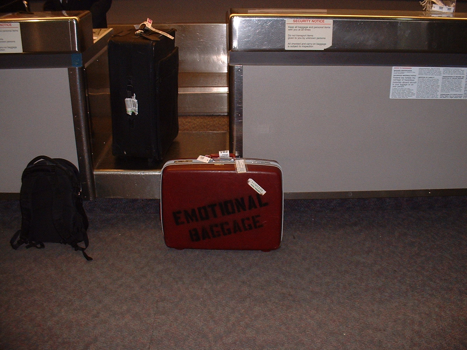Image: Retro red suitcase with 'Emotional Baggage' stenciled on side at checkin counter in front of scale.