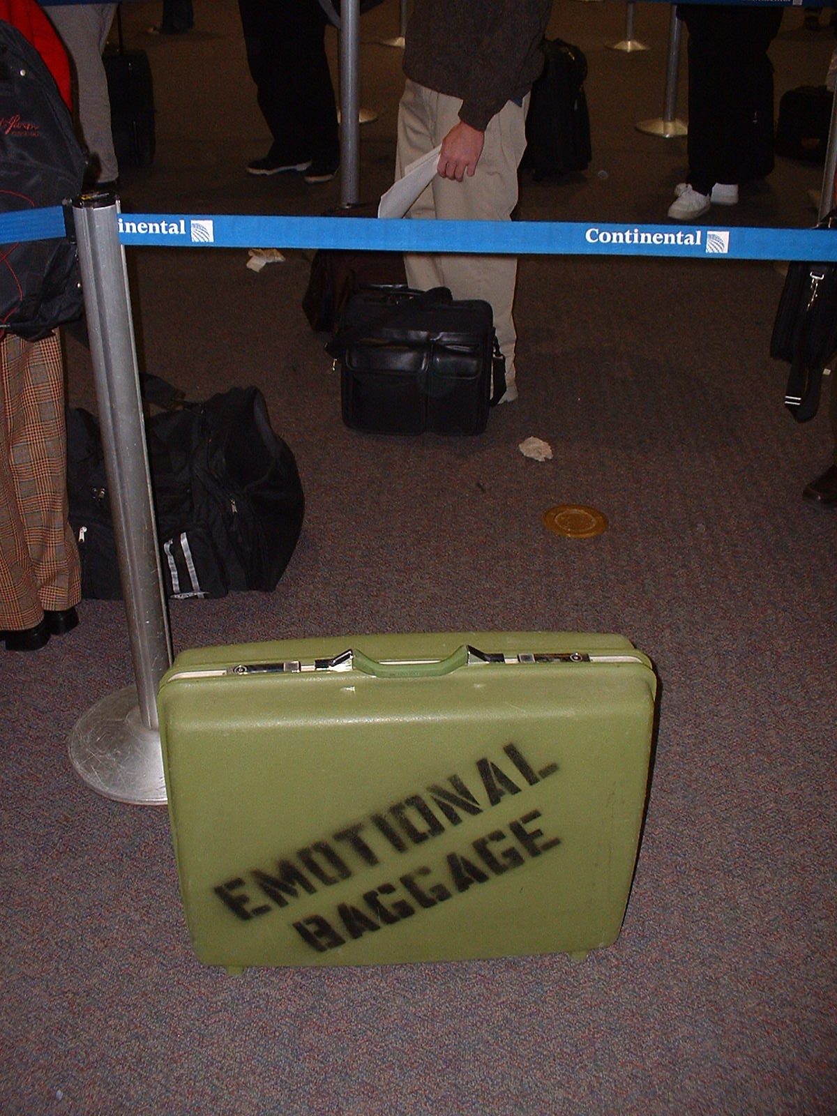 Image: Retro green suitcase with 'Emotional Baggage' stenciled on side in check-in line.