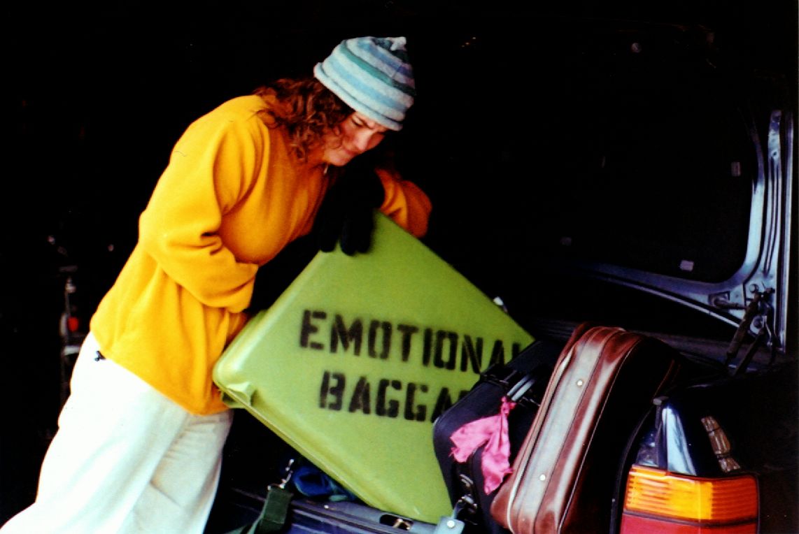 Image: Retro green suitcase with 'Emotional Baggage' stenciled on side with woman putting it in trunk.